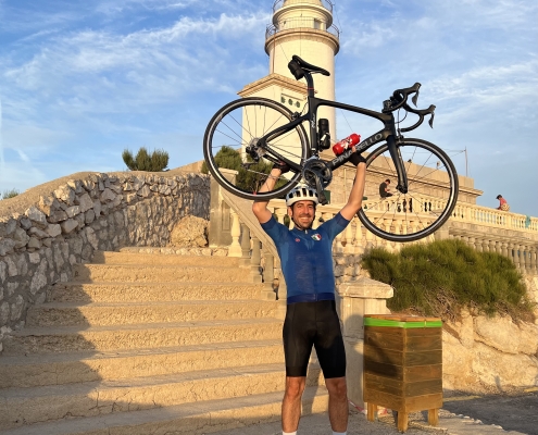 Dr. Roberto Vélez levantando su bicicleta frente a un faro al atardecer, simbolizando la importancia del ejercicio en el tratamiento del cáncer.
